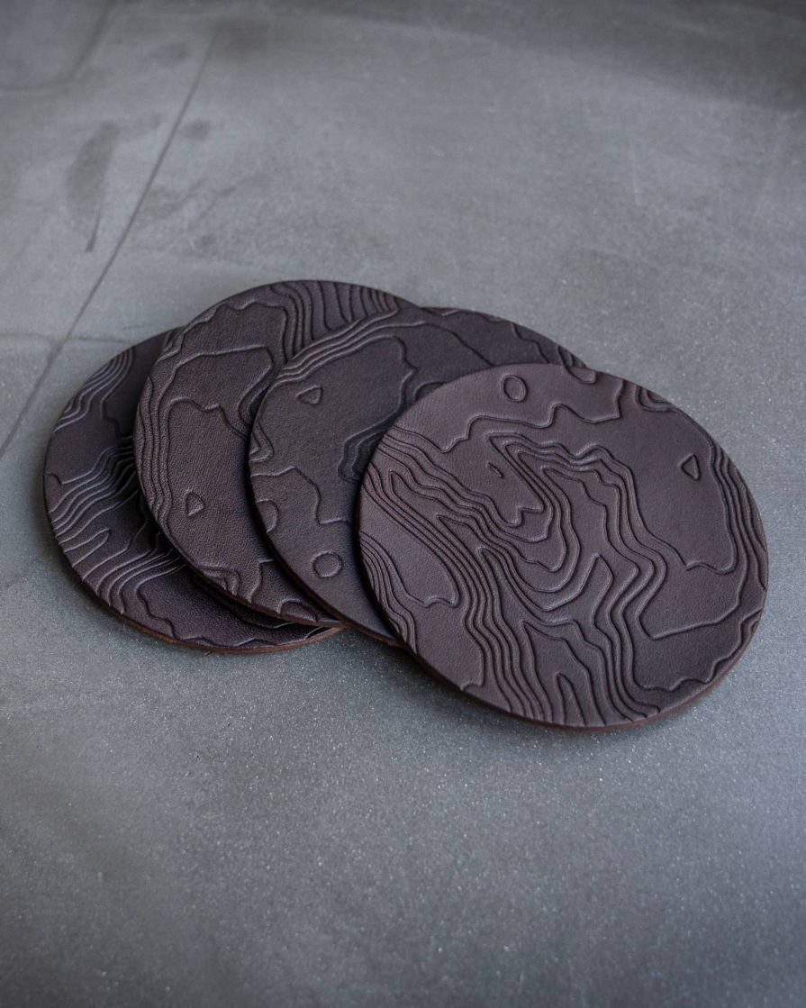 Dark brown leather coasters featuring a topographic pattern debossed on the surface.